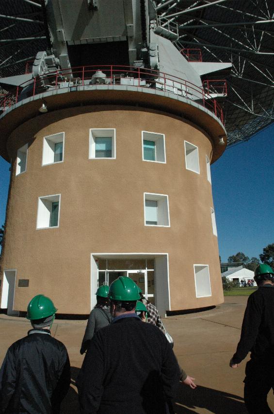 People with green hard hats in front of circular brown building below telescope dish