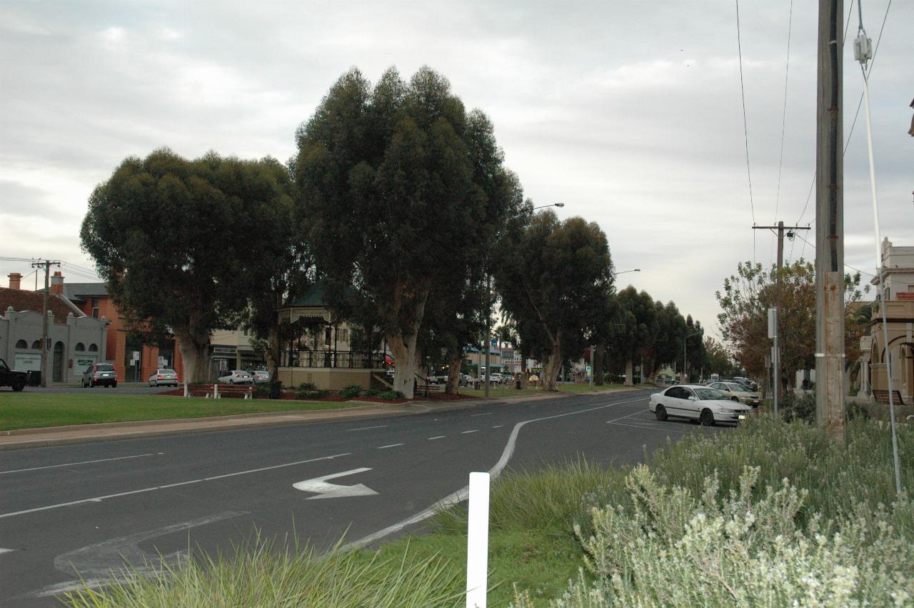Looking down median strip with trees and grass, and a rotunda in among the trees, cars parked on the outside of the roads