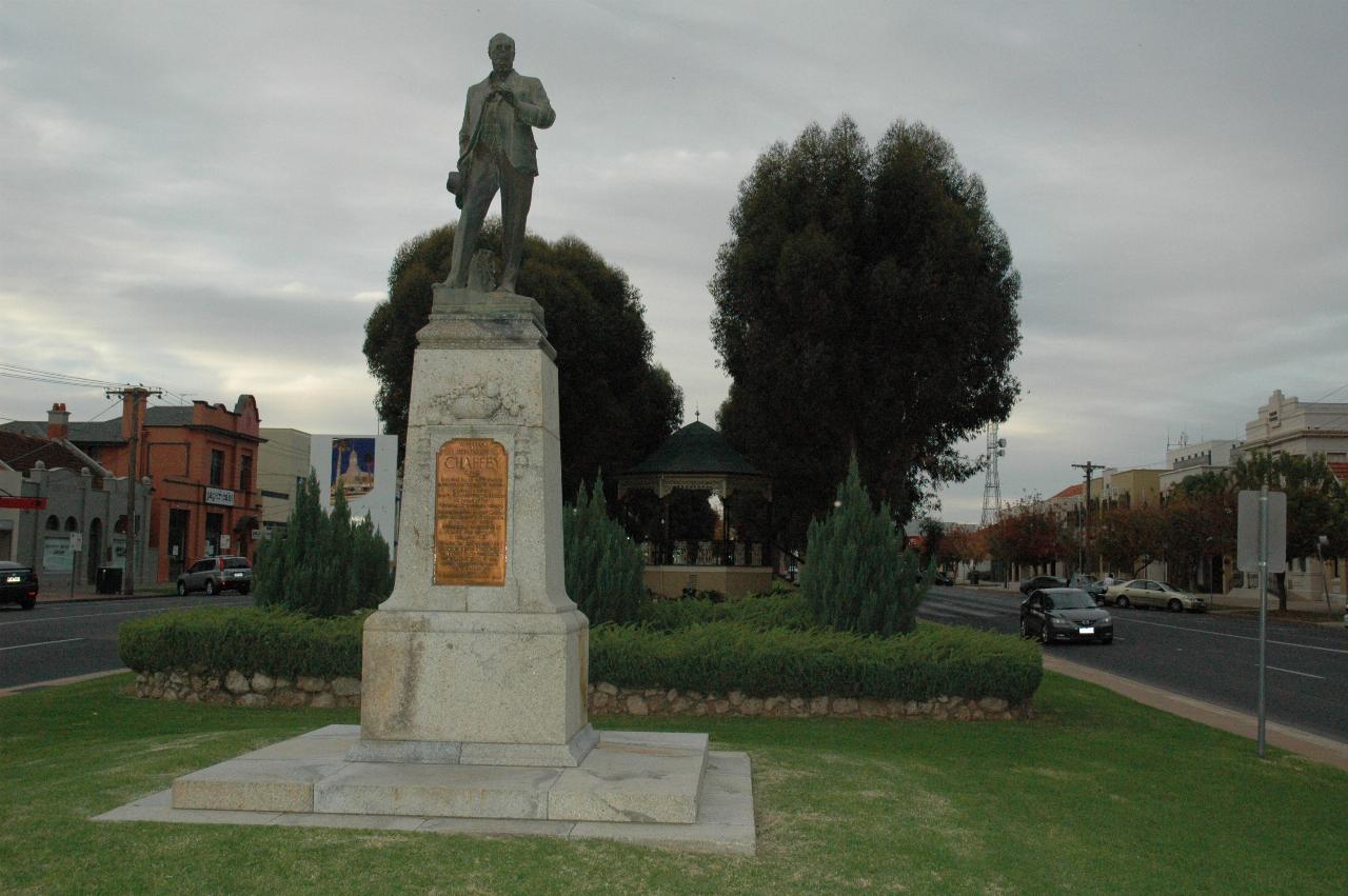 Statue of man with inscription, rotunda and trees behind, all in the median strip