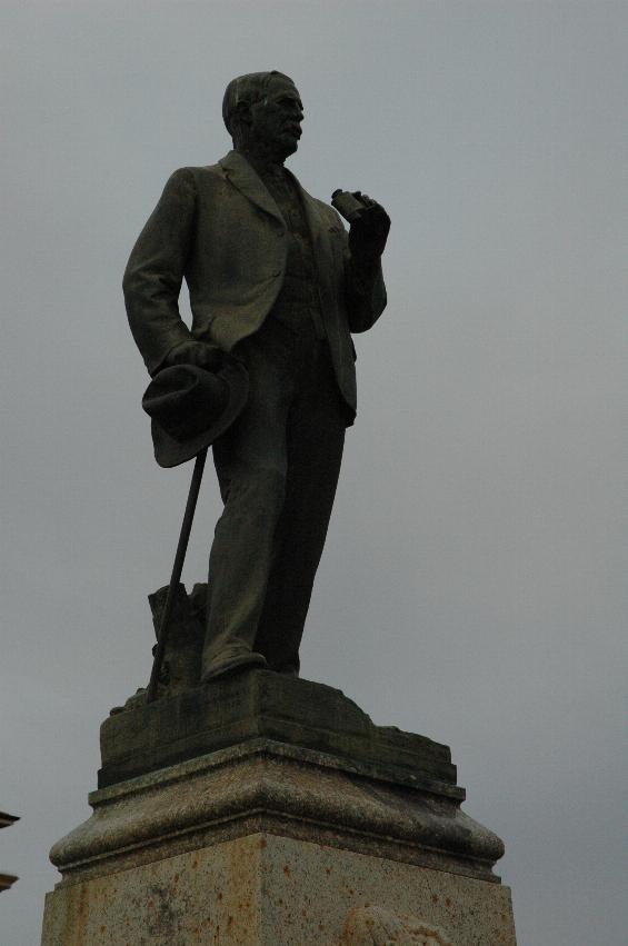 Close up of man on pedestal, holding binoculars in right hand, walking stick and hat in left