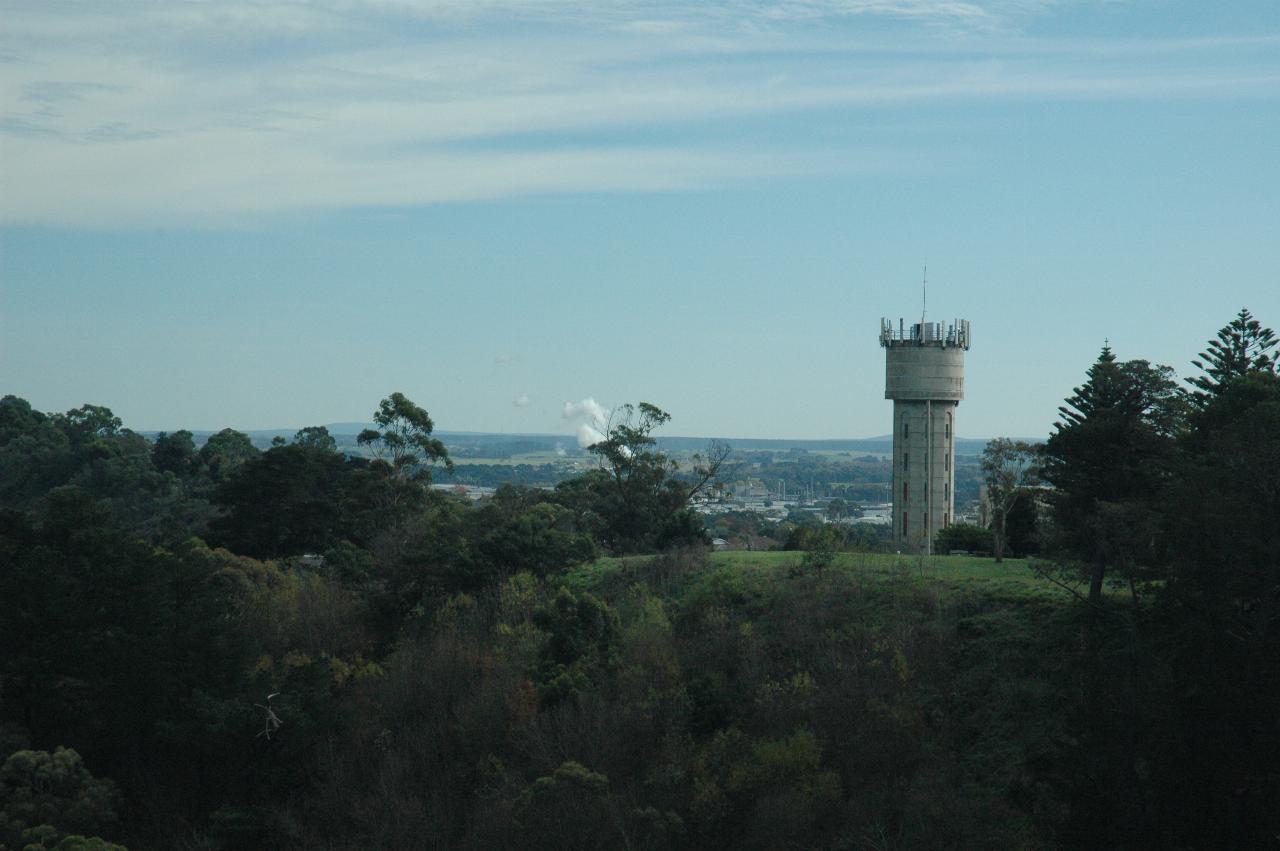 View to cement water tower on grassy hill, some buildings and rising steam cloud in distance
