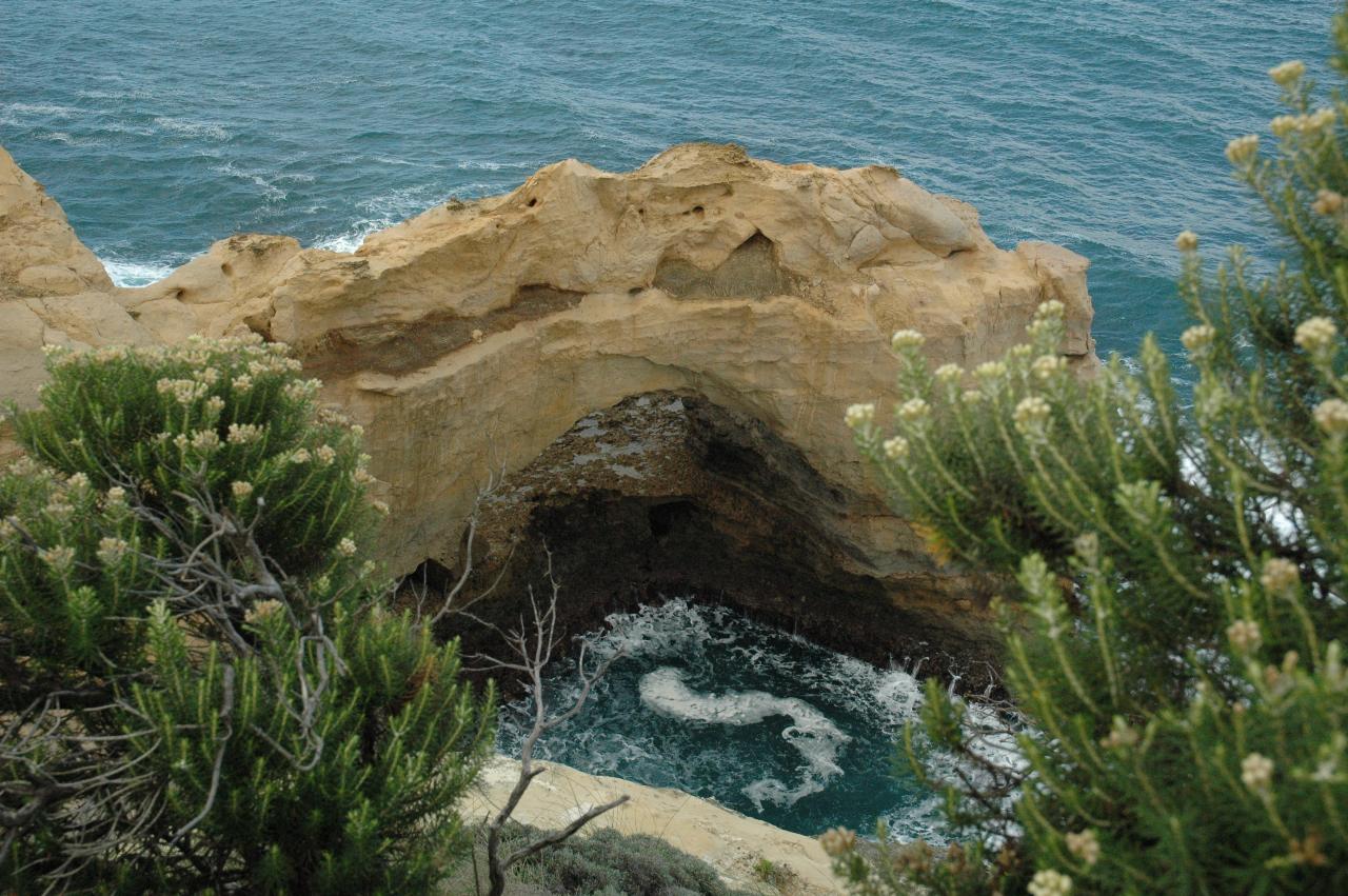 Natural arch of ochre coloured material, one end in ocean with white water around the base and vegetation in foreground