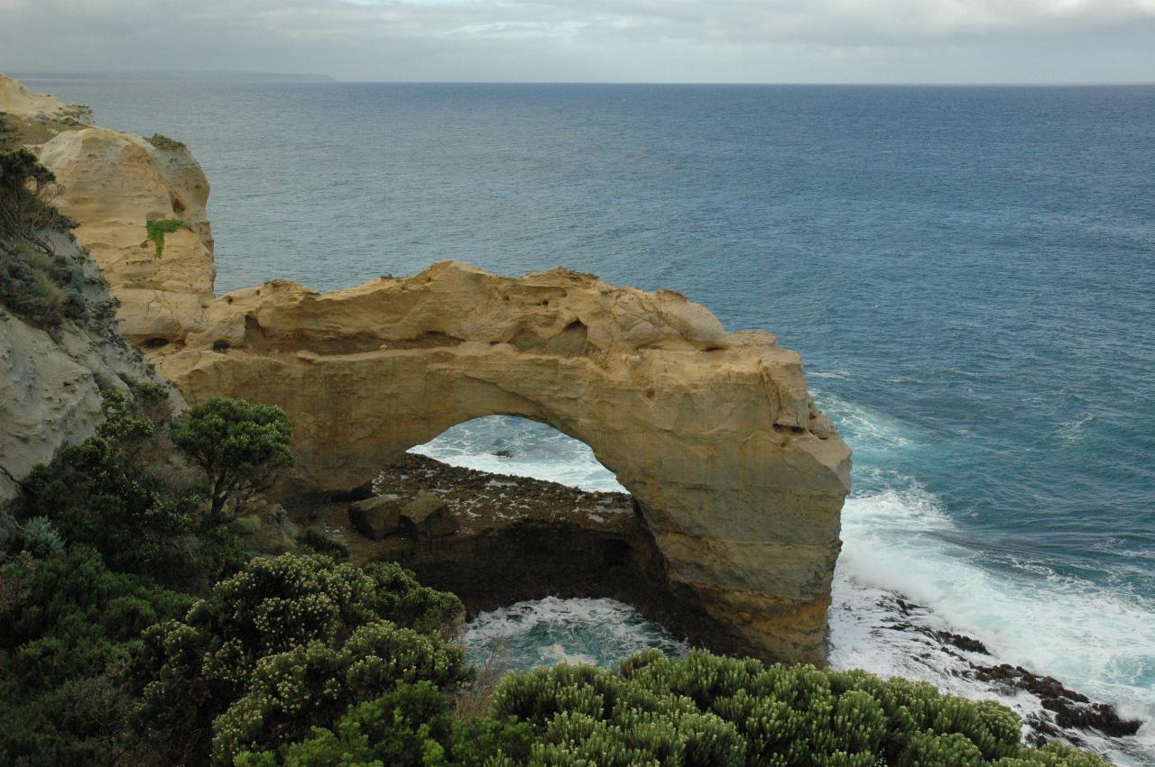 Natural arch of ochre coloured material, one end in ocean with white water around the base