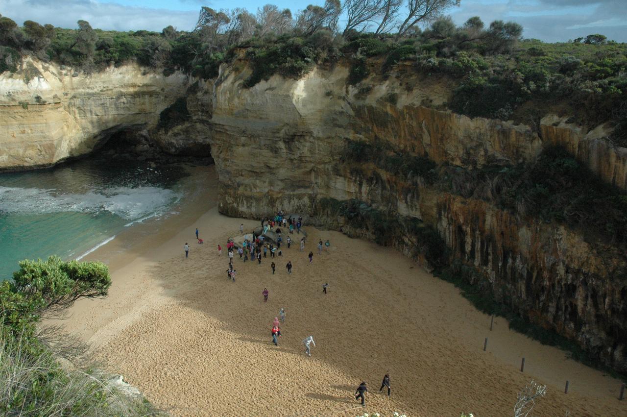 Small looking people on sandy area at base of tall cliffs, with a wave on the beach