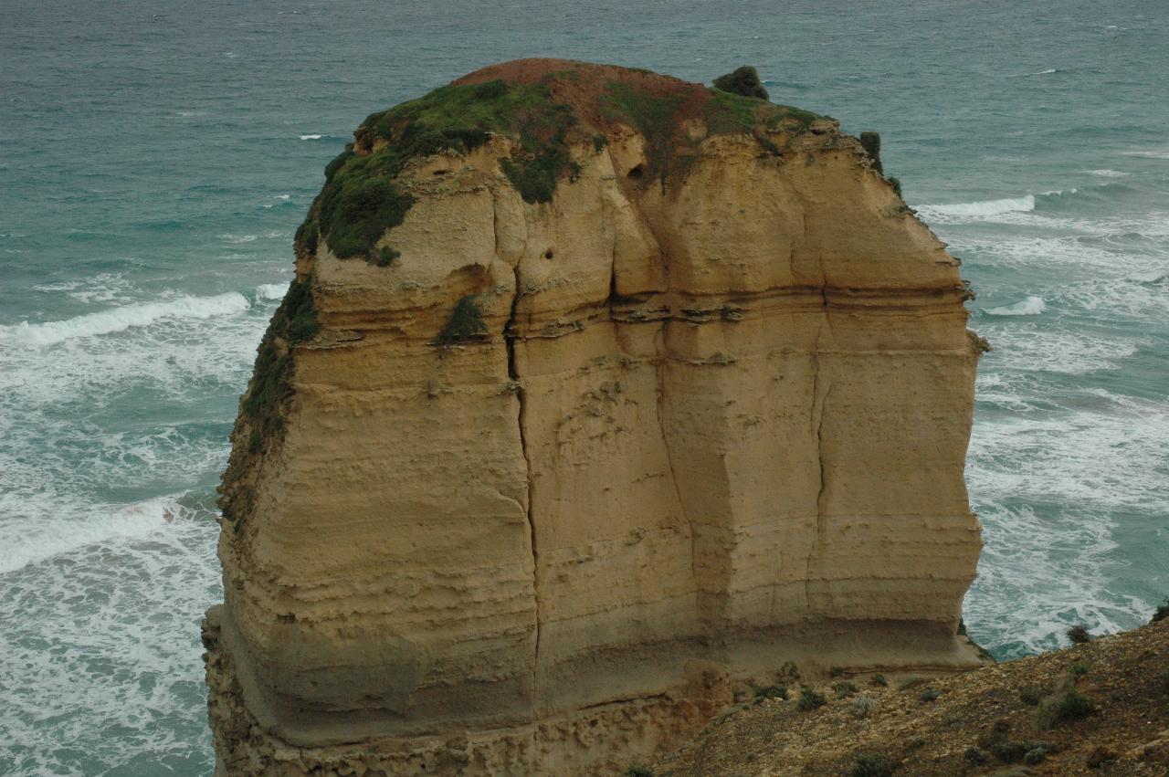 Ocean with waves as backdrop to pillar of rock with vegetation on top, and big crack down the front