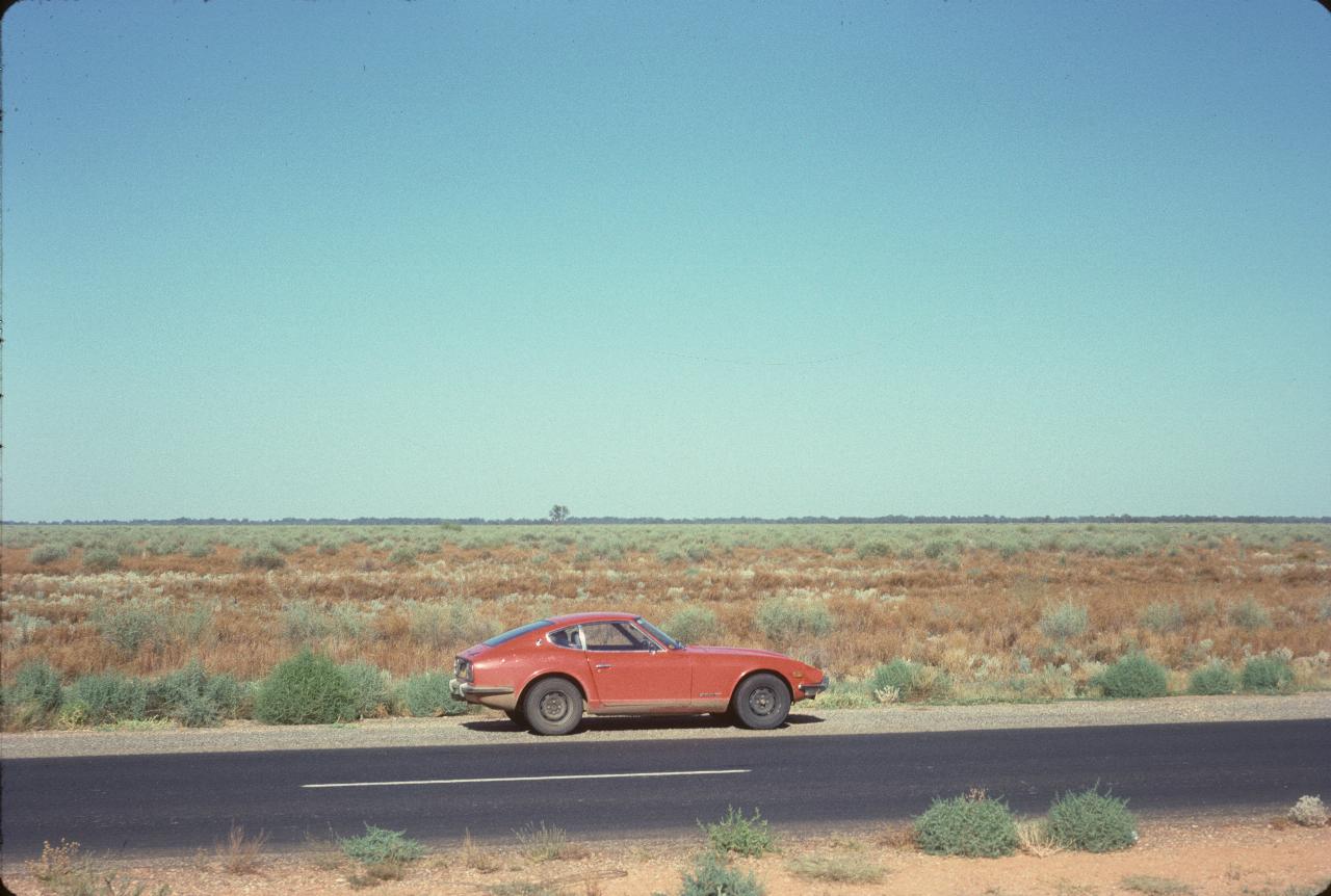 Red car parked on the side of the road, flat country to horrizon, with low vegetation