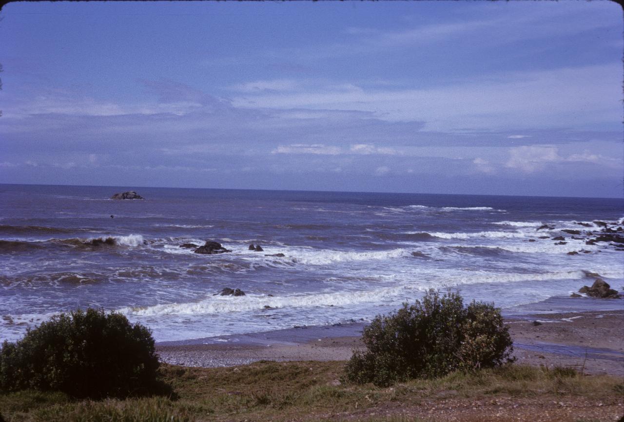 Sea shore with brownish water, small waves breaking and some low rock formations in the water