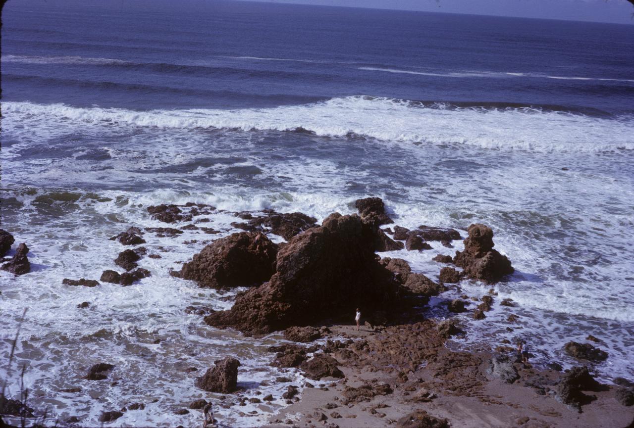 Looking down on dark brown rock formation at edge of sea with person nearby