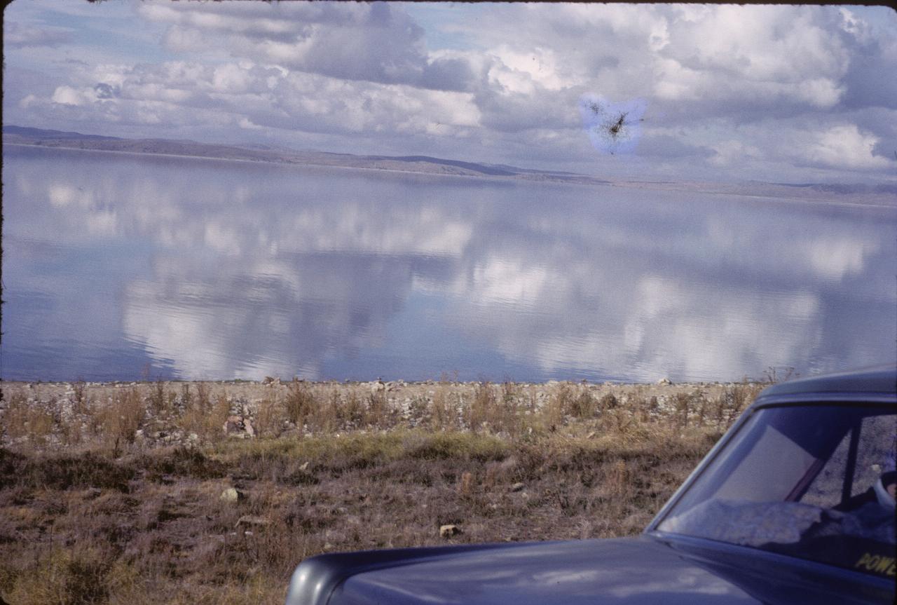 Car parked on verge of calm lake reflecting clouds