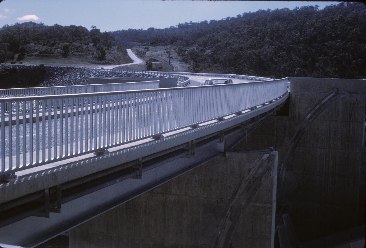 Car passing over concrete bridge atop spillway, road heading up hill