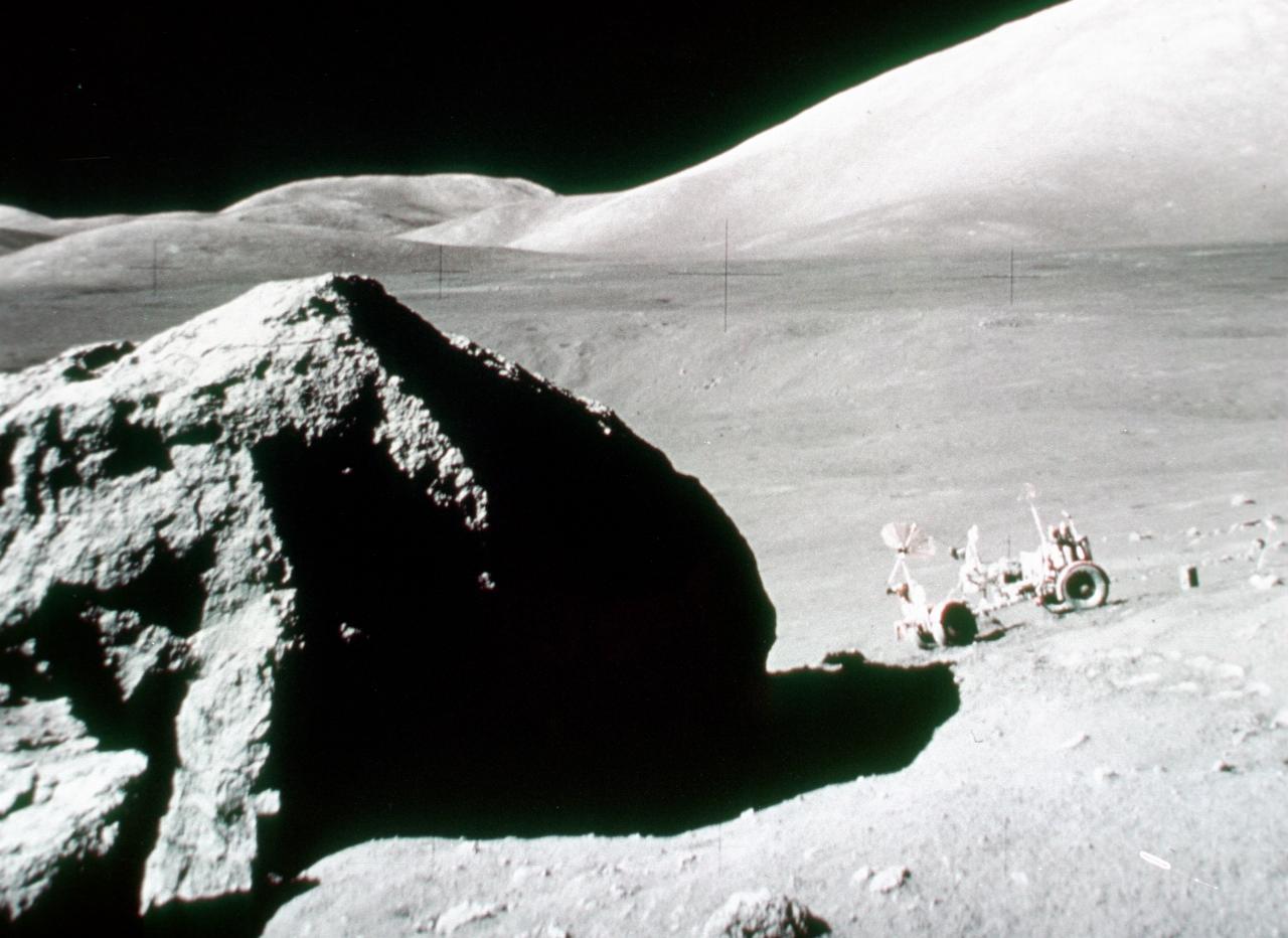 Lunar rock in foreground, rover parked some distance on downhill slope