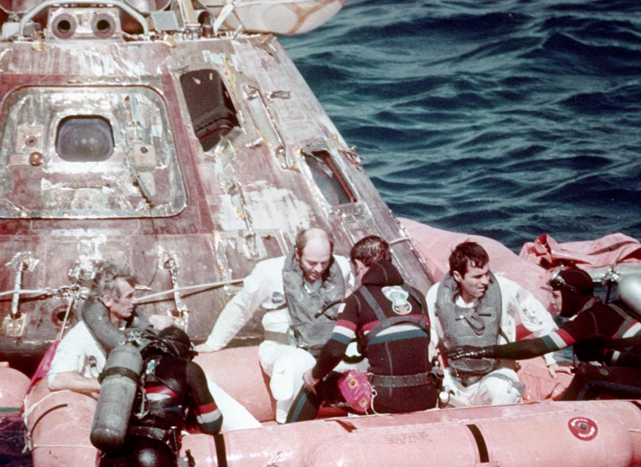 Astronauts and divers in rubber dinghy next to capsule in water