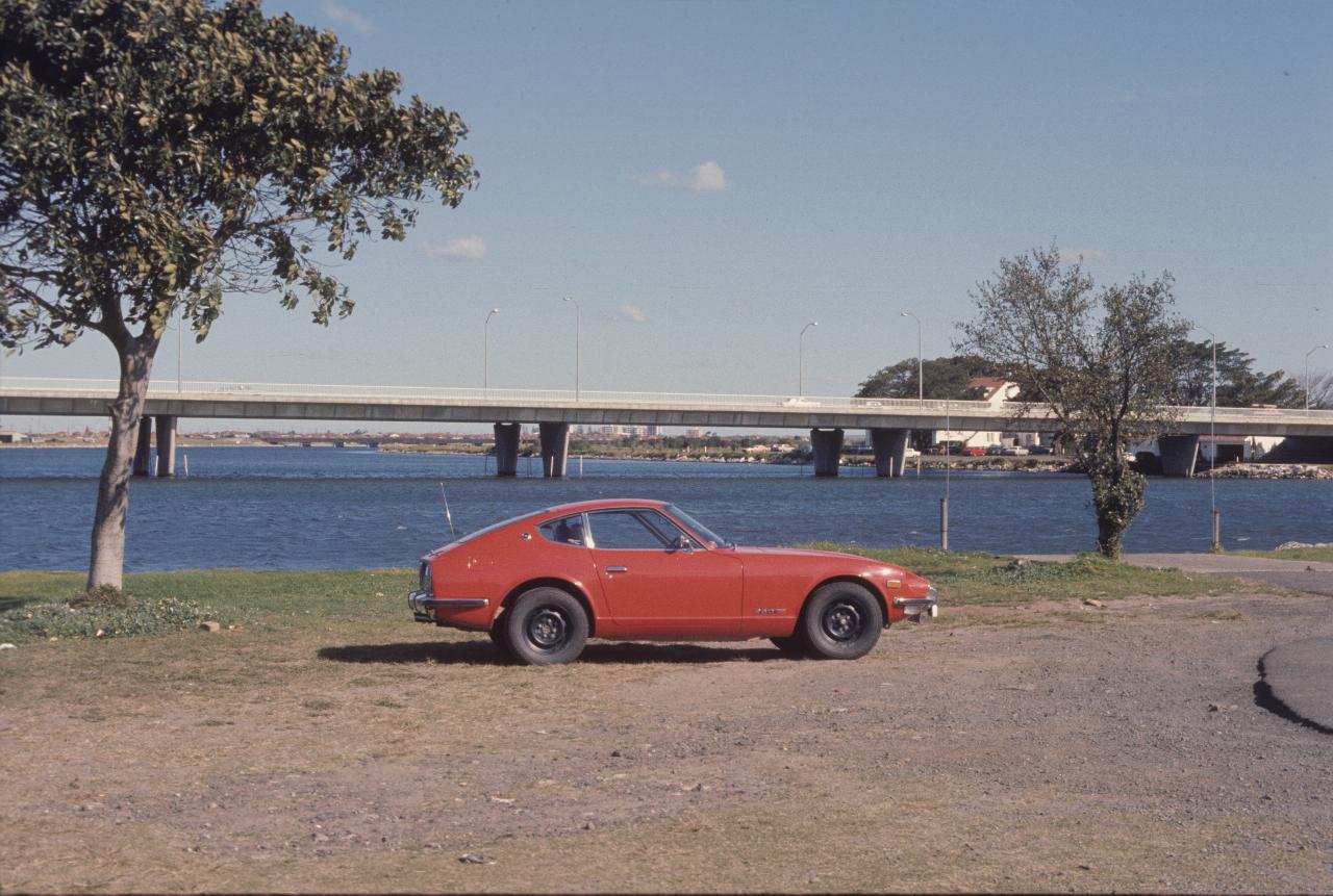 Red car on river bank, bridge crossing river behind