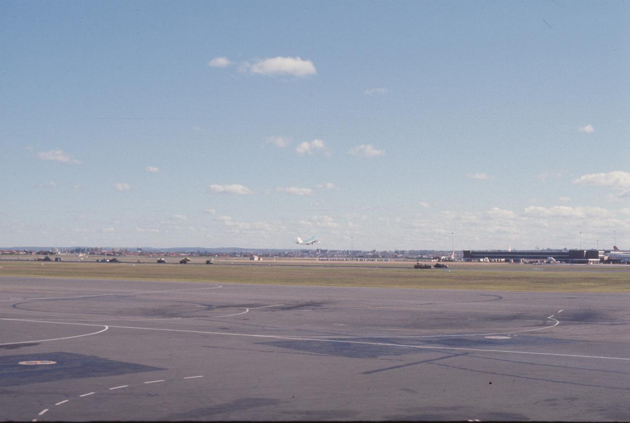 View across airport; plane taking off; runway under repair and terminal in the distance