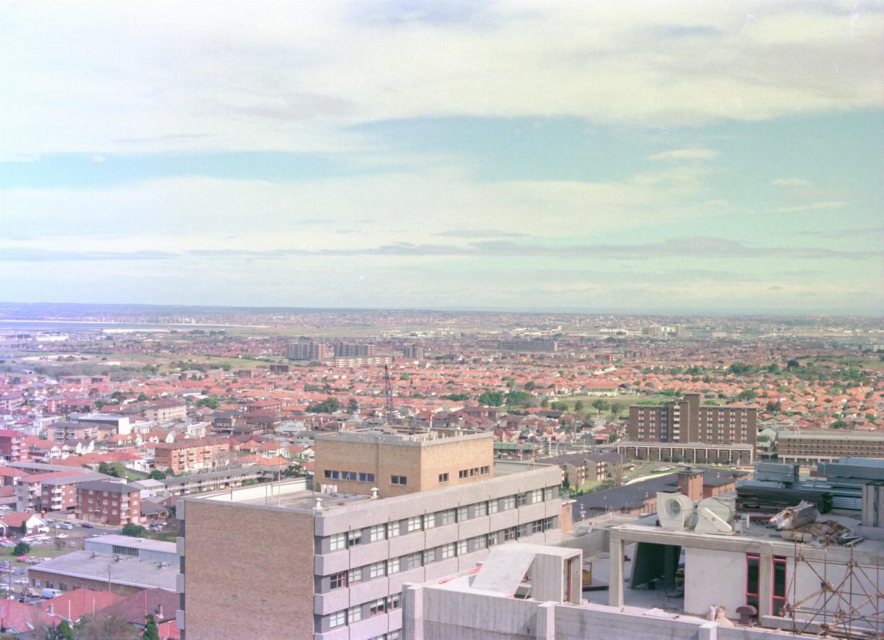 Nearby tall buildings across sea of red roofs to airport and more suburbia, and a bay