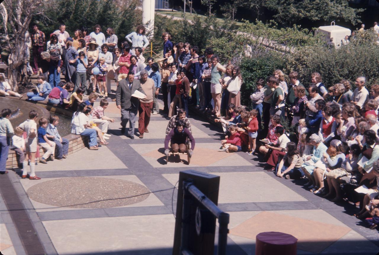 Billy cart style vehicle coming around fountain in front of crowd