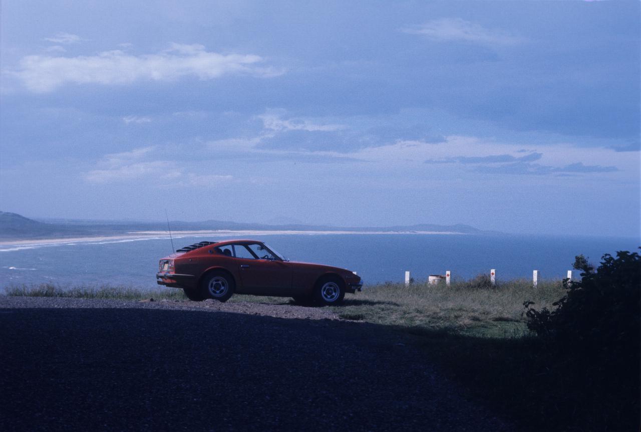 Car on headland, with sandy beach, breaking waves and distant headland beyond