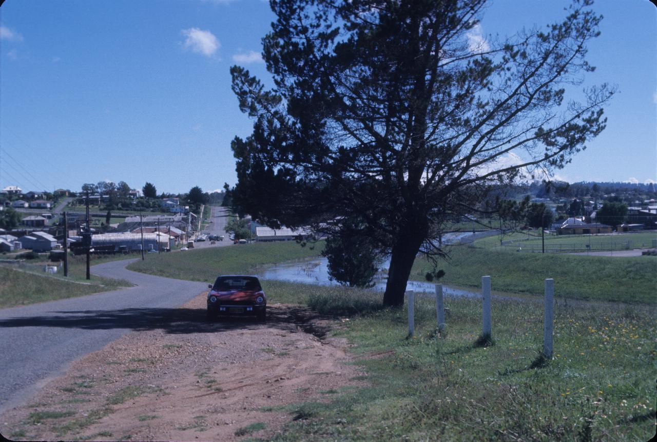 Car parked in shade of tree, next to river with small town behind