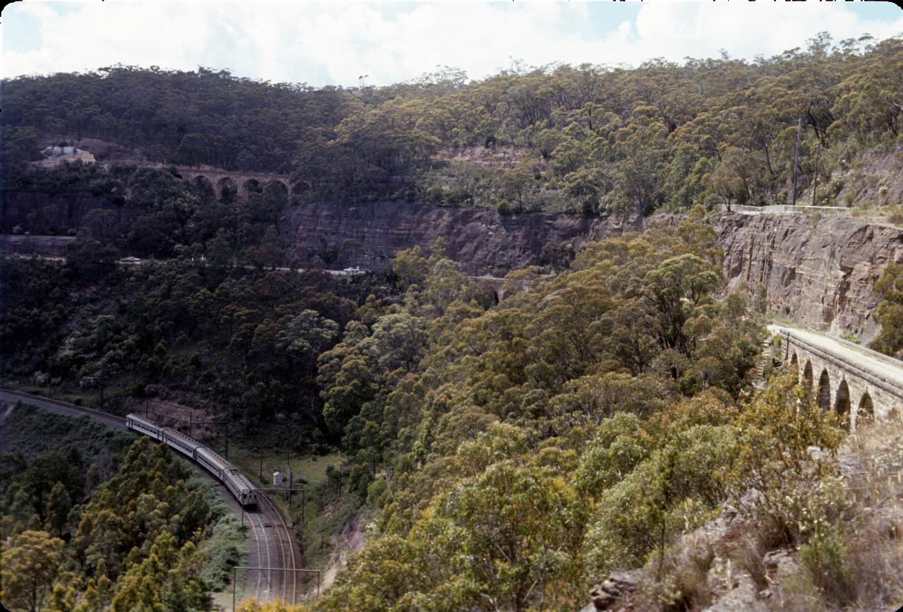 Train on tracks at base of valley with stone bridges on side