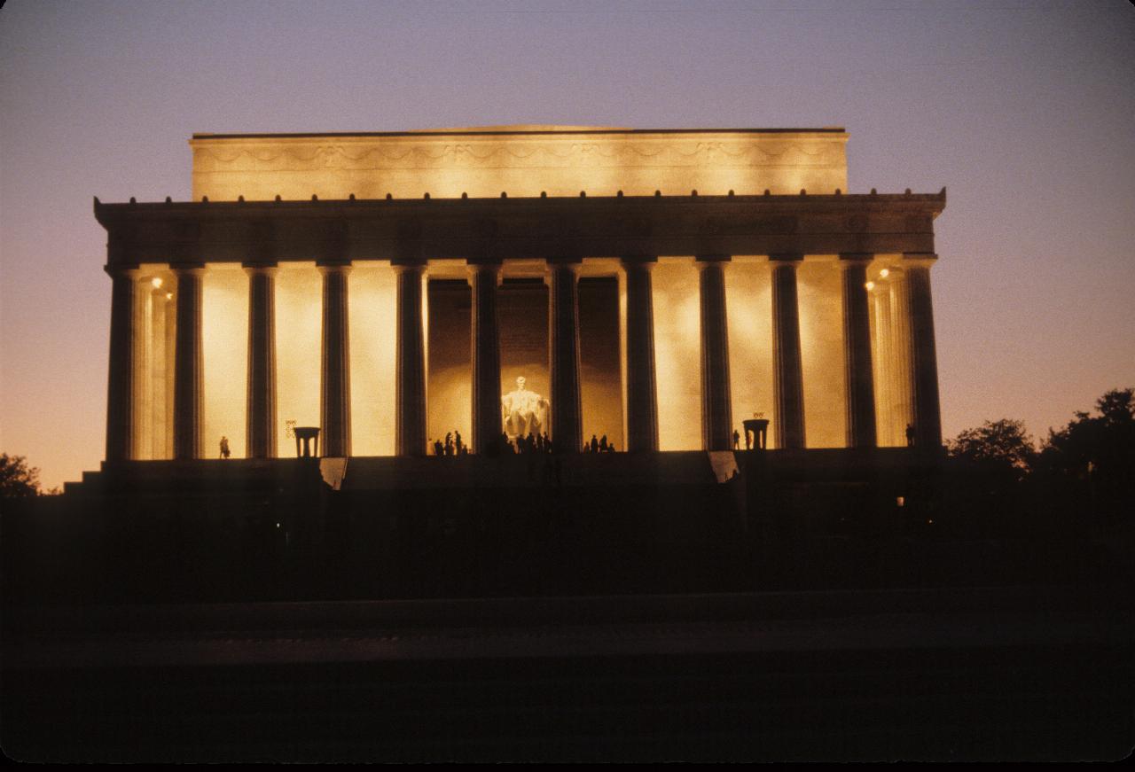 Large building, illuminated with front pillars in shilouette, big statue inside. people around