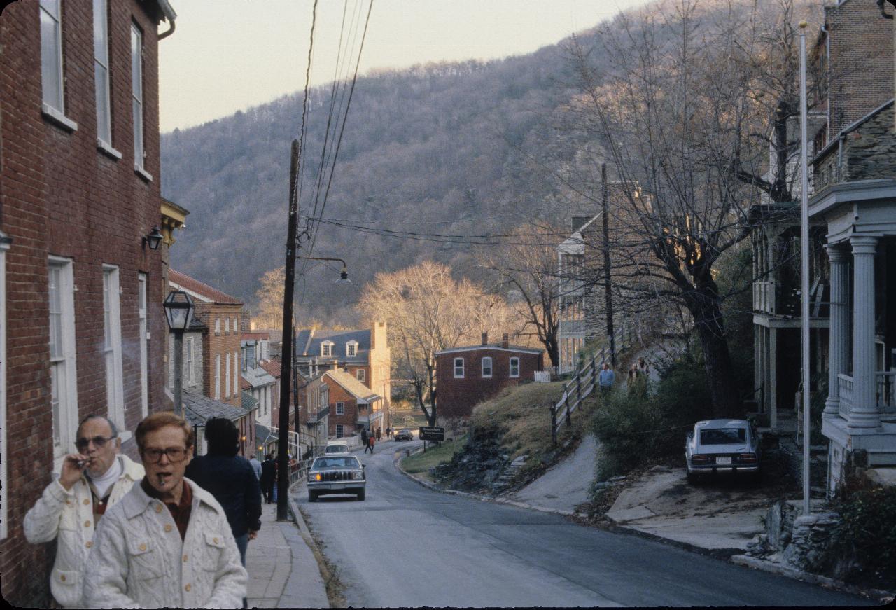 Street scene, with houses on both sides,a couple of cars and a few people on narrow footpath