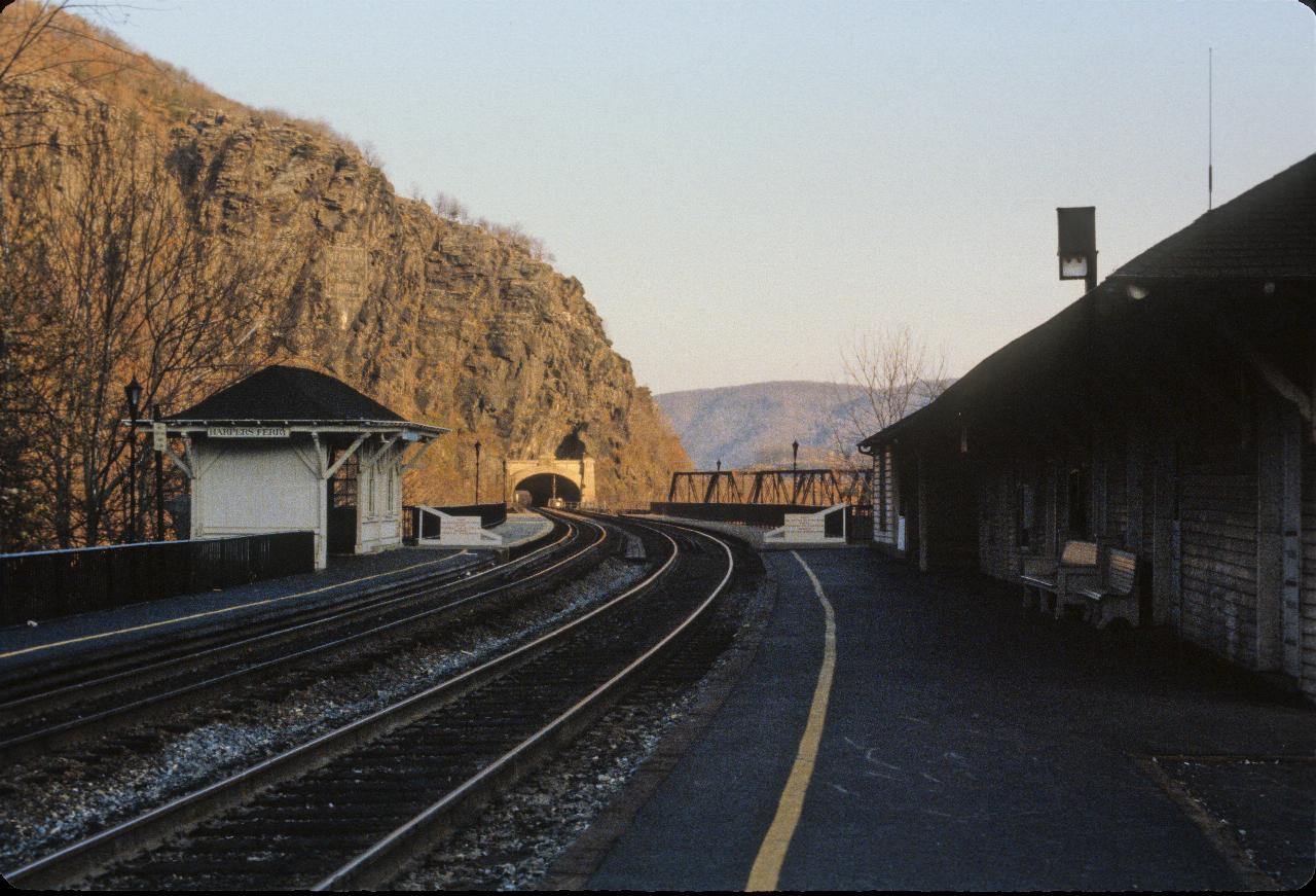 Dual rail tracks curve, cross bridge and enter tunnel, with a platform on both tracks