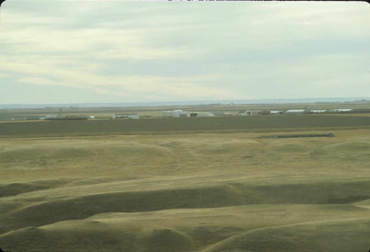 Coulee in foreground, to plowed field with a line of buildings behind to mountains in far distance