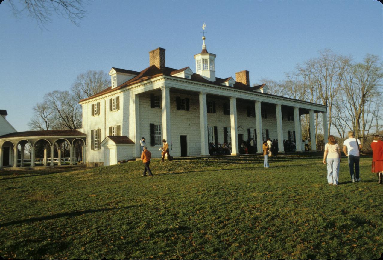 White, two storey house with red roof, large porch with white pillars, leading to grassy area