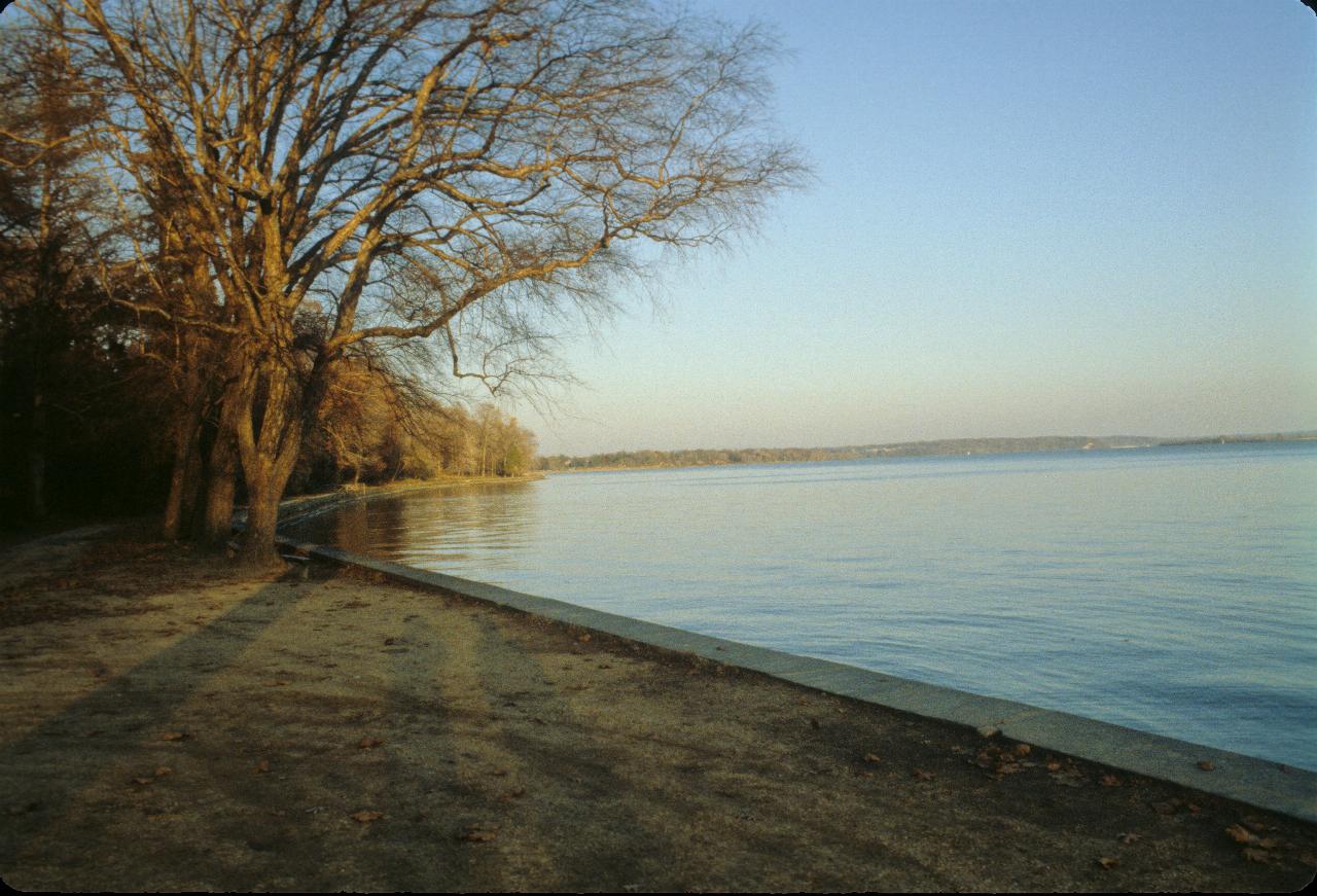 Large expanse of water, distant trees on far bank, and walkway and trees on this river bank.