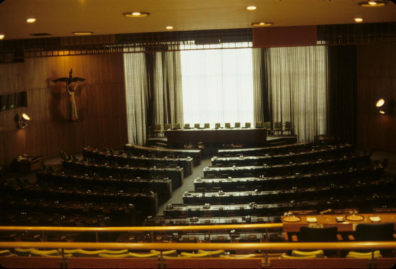 Conference room with straight table on a raised up section at rear with a window letting in the light; rows of seats in front