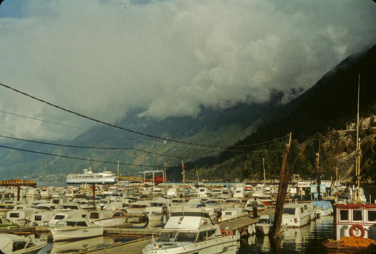 A mass of moored pleasure boats, with car ferry further back and steep, tree covered hills behind