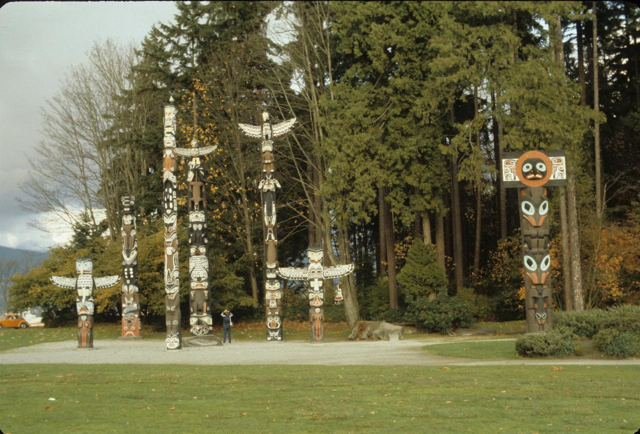 Seven totem poles in front of a stand of mostly evergreen trees