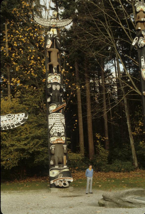 Man standing next to totem pole, looks small, with trees behind