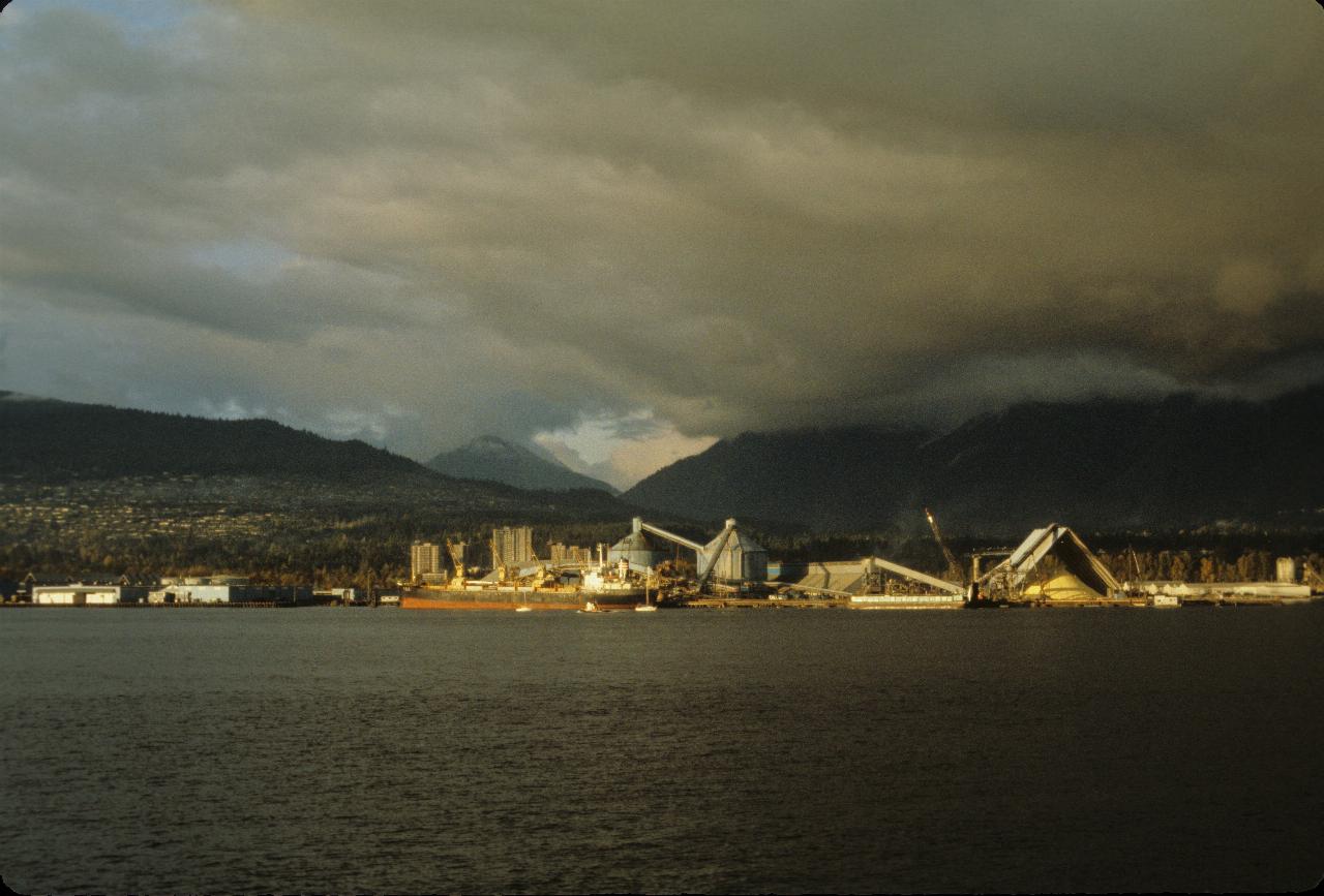 Across water to ship dock near loader, piles of sulphur and mountains behind