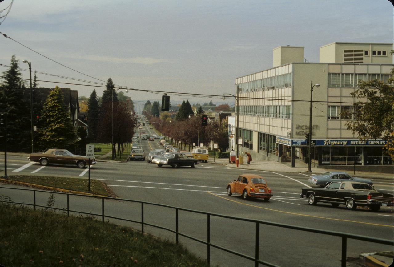 Street scene at an intersection; cars waiting at traffic lights, small office building on the corner.