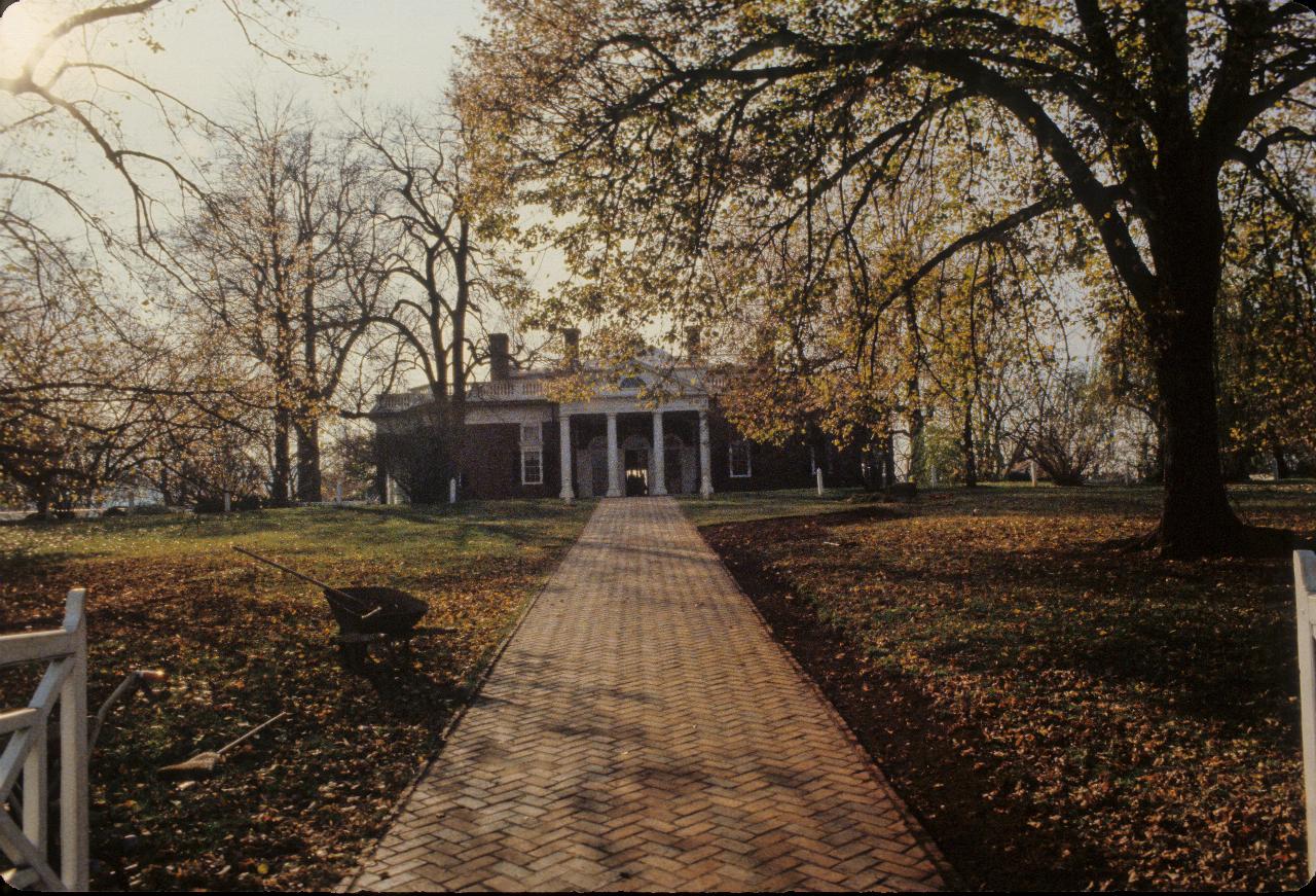 Brick path leading to red brick house with white pillars in front, partly hidden by trees in autumn colours