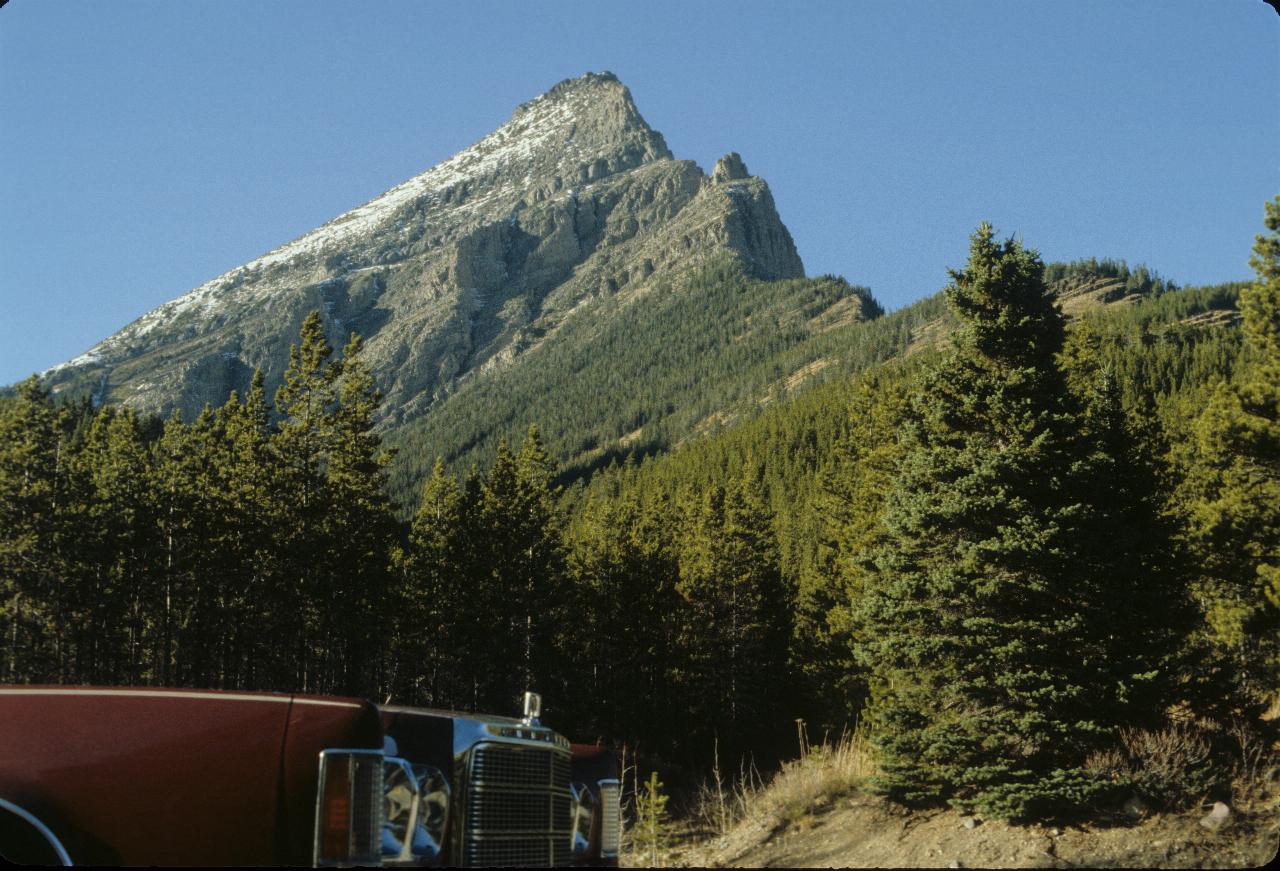 Front of car, pine trees running part way up craggy, triangular shaped hill with a dash of snow on the top