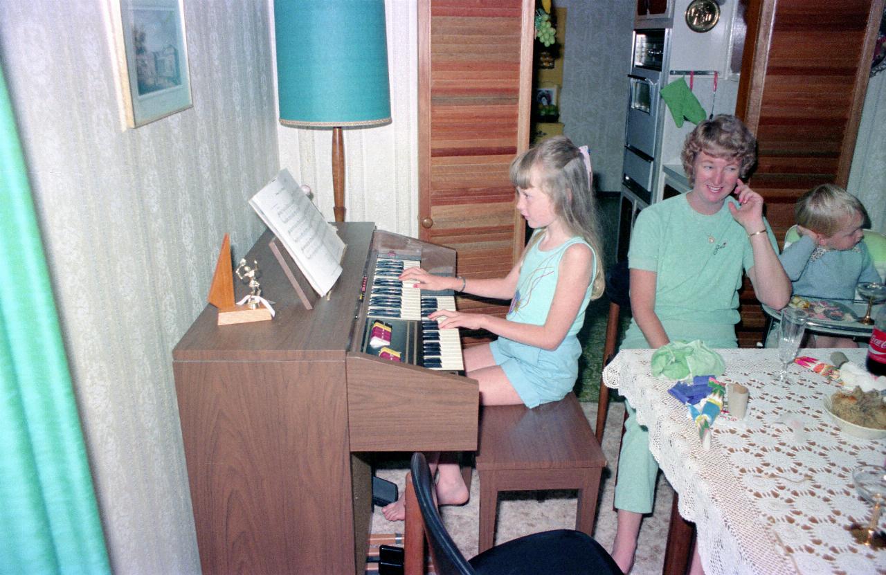 Young girl playing organ; mother and brother sitting nearby at table