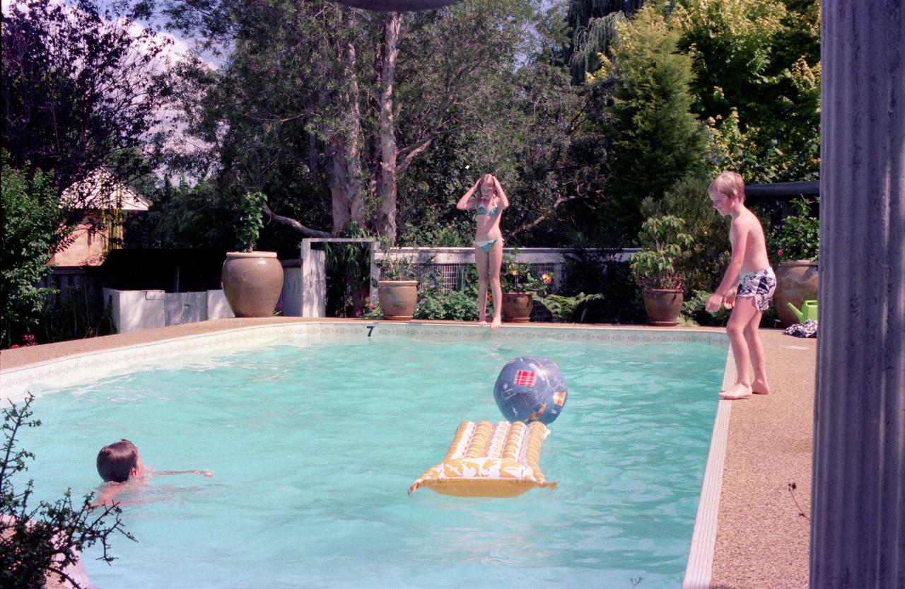 Swimming pool with young boy in, another about to jump from side, and girl at far end about to dive in