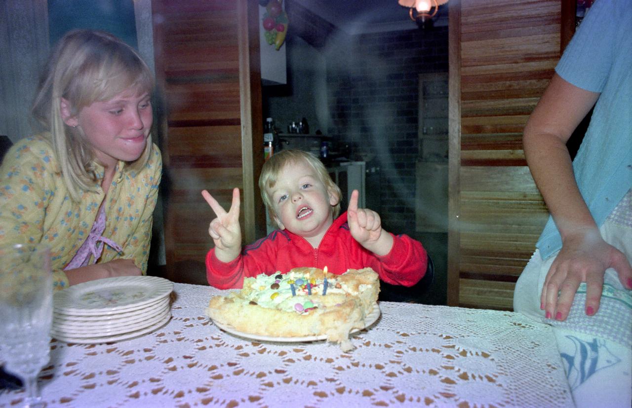 Little boy with three fingers with cake and 3 candles on table in front