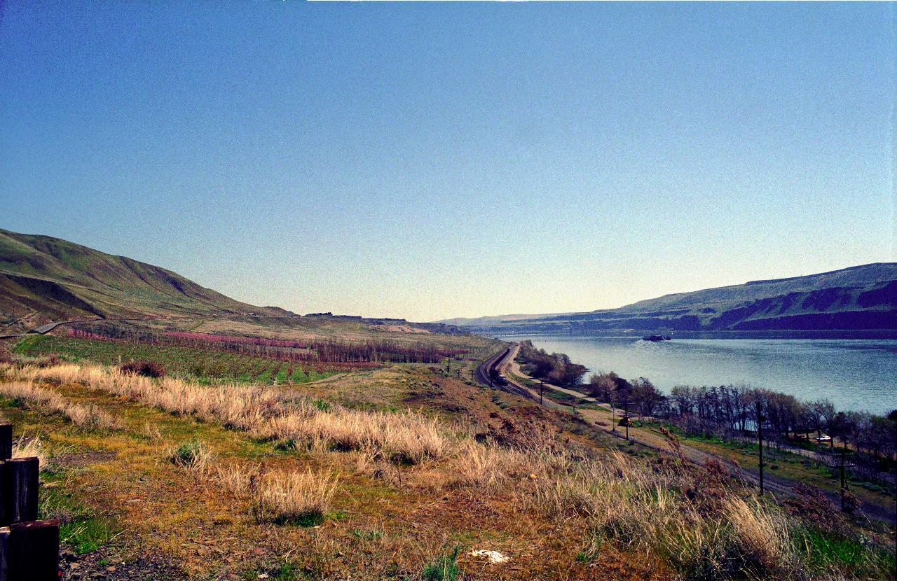 Broad river with barges between low cliffs and railway line this side