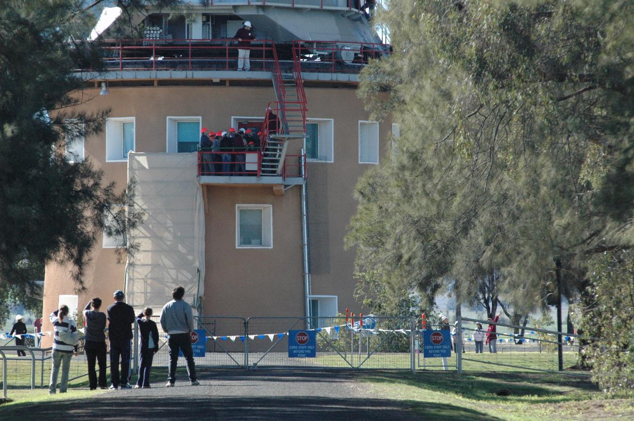 Side of circular building with people climbing stairs on outside to reach base of telescope mechanism