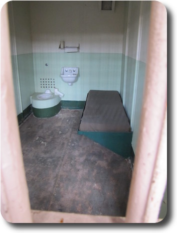 View through bars to spartan cell - bed, toilet, basin