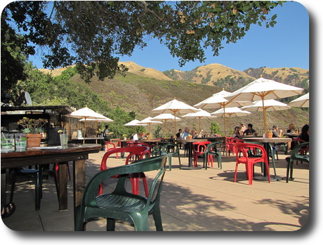 Eating area with tables with umbrellas, and coastal, mostly brown hills behind