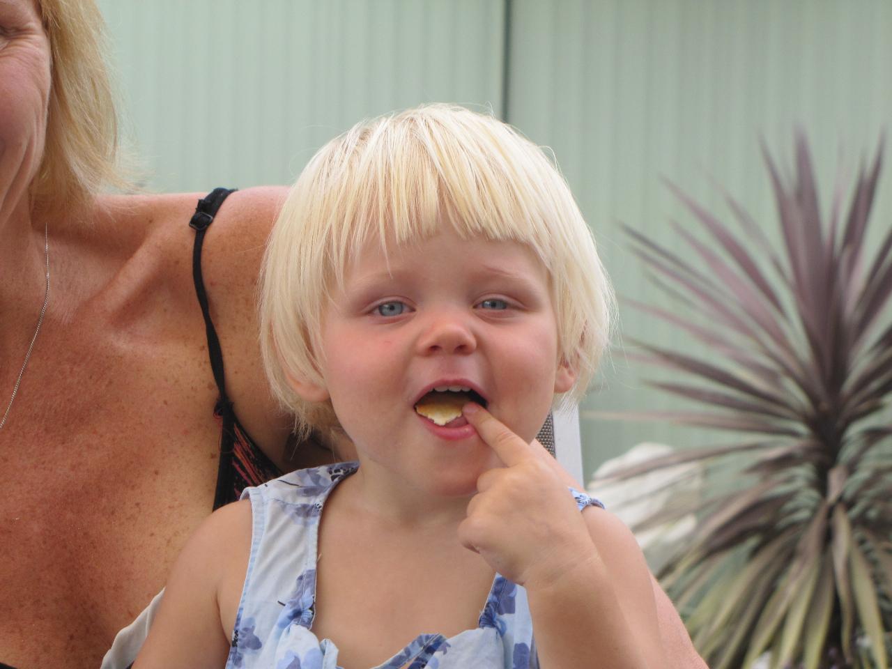 Blonde haired little girl with potato chip in mounth, on mum's lap