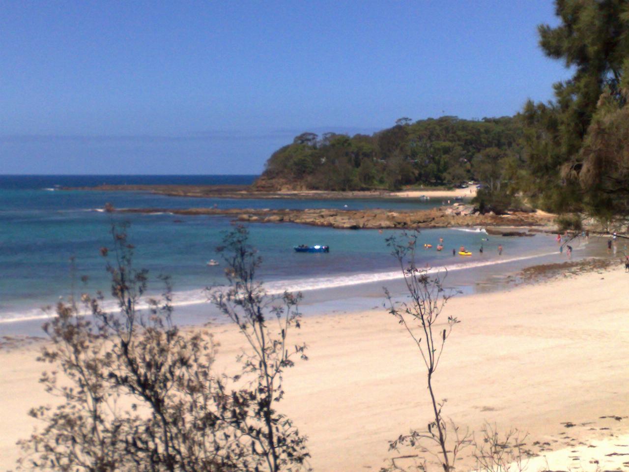 Sandy beach with a nearby, tree covered headland; boats and people on the beach