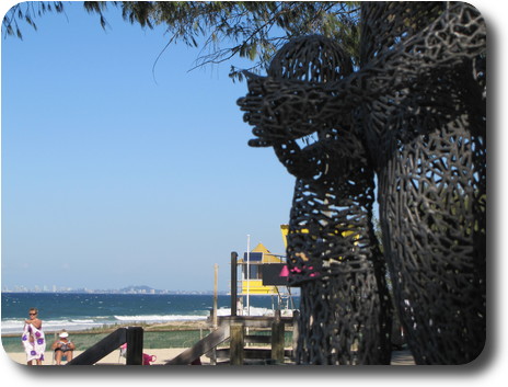 Distant high rise buildings, nearby beach and part of sculpture with woman's hands on child