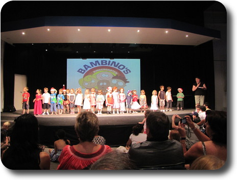 About 25 children in a line across the stage; supervisor m/c on right