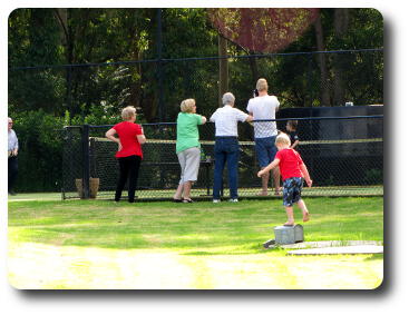 Little boy in foreground, man and boy on tennis court, 4 adults with camera on side of court