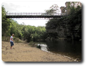 Man standing on sand beside river, below suspension bridge